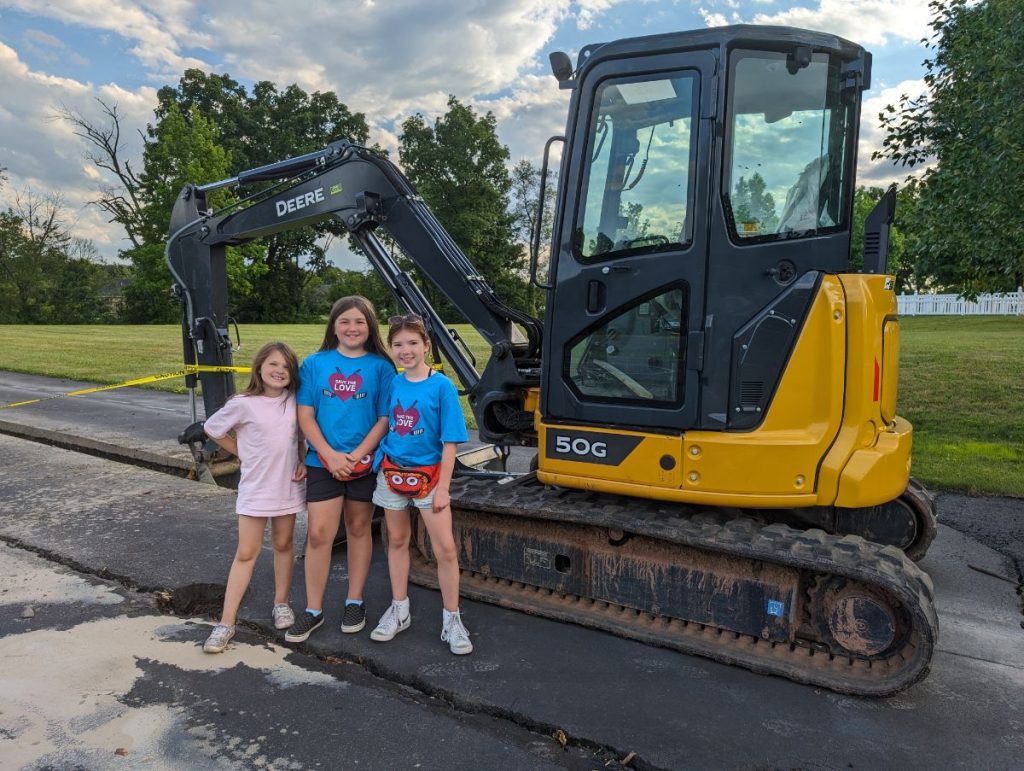Natalie Van Druff, Lilly Walter, and Olivia Van Druff stand with a construction vehicle while their community's outdoor dek hockey rink is being fixed up!